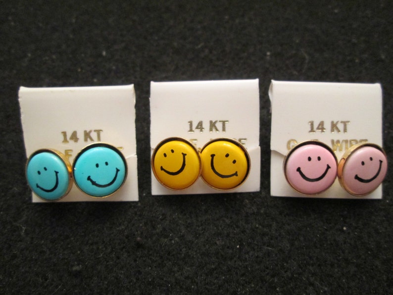 May include: Three pairs of 14KT gold smiley face earrings. The earrings are in blue, yellow, and pink.