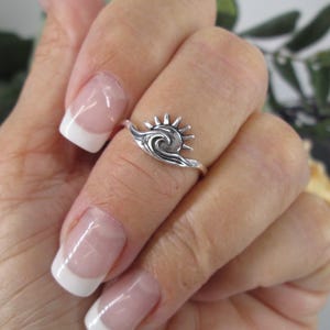 May include: A silver ring featuring a sun and wave design. The ring is worn on a finger, showcasing the intricate details of the sun's rays and the flowing wave. The ring is a piece of jewelry.