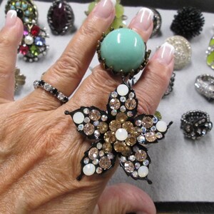 May include: A hand wearing a variety of rings, including a large ring with a turquoise stone and a flower-shaped ring with rhinestones.