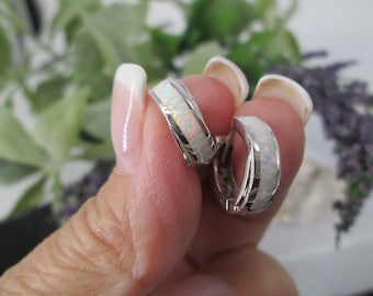 May include: A pair of silver hoop earrings with a white opal inlay. The earrings are held up by a person's fingers, with a blurred background of greenery and purple flowers.