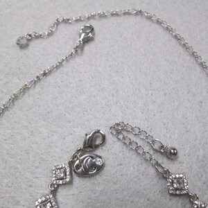 May include: A silver necklace with a silver charm featuring a small crystal and a logo. The necklace has a chain with a lobster clasp and an extender chain.