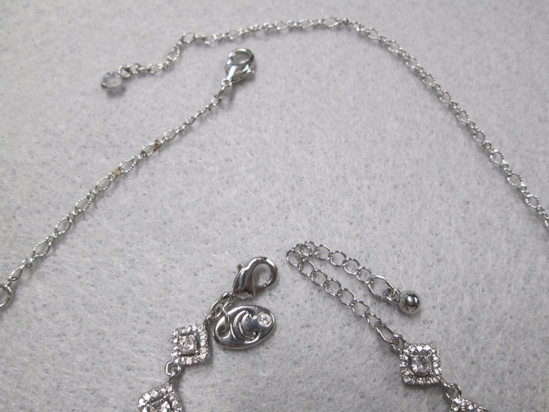 May include: A silver necklace with a silver charm featuring a small crystal and a logo. The necklace has a chain with a lobster clasp and an extender chain.