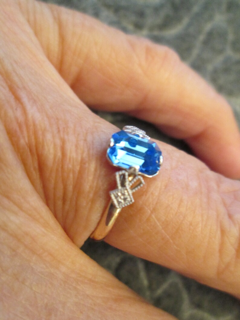 May include: A gold ring with a blue gemstone and white accents. The ring is on a finger.