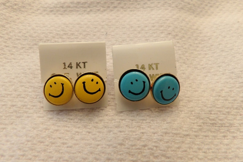 May include: Two pairs of 14KT gold smiley face earrings. One pair is yellow and the other is blue.