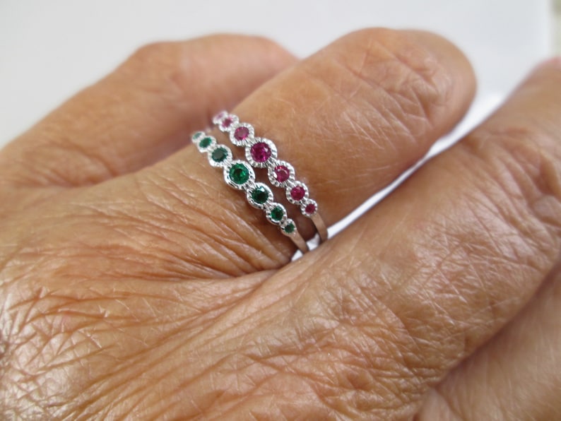 Pu&ograve; includere: Due anelli d'argento con piccole pietre preziose verdi e rosse. Gli anelli sono impilati su un dito.