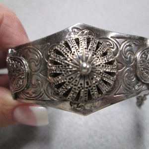 May include: A silver bracelet with an ornate design and a domed center piece. The bracelet has a chain attached to one end.