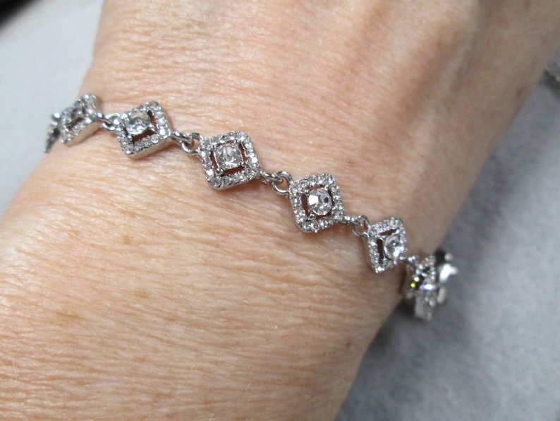 May include: A silver bracelet with a row of square-shaped crystal accents. Each square has a smaller crystal in the center.