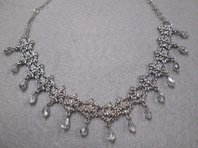 May include: A silver necklace with a delicate chain and a floral design. The necklace features dangling crystal drops.