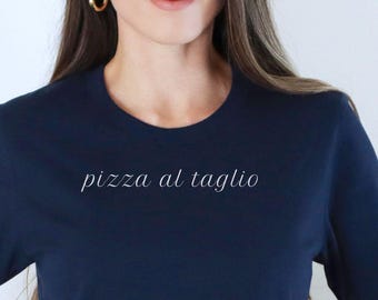 May include: Navy blue t-shirt with the text "pizza al taglio" in a cursive font. The shirt has a classic crew neck and short sleeves. The model is wearing blue jeans and a brown belt.