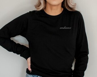 May include: Black long-sleeve shirt with the word "andiamo" in cursive script on the upper left chest. The shirt is paired with light blue denim jeans. The model is standing in front of a plain, light gray background.