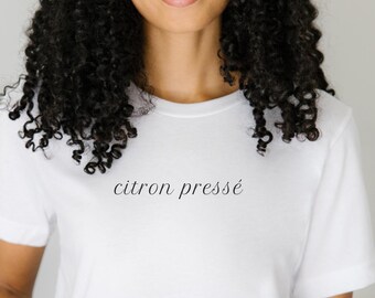 May include: A white t-shirt with the phrase "citron pressé" in a delicate, cursive font. The shirt has short sleeves and a crew neck. The model is wearing blue jeans.