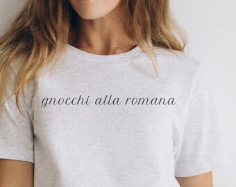 May include: A light gray t-shirt with the text "gnocchi alla romana" in a cursive font. The shirt is paired with black pants. A black watch is visible on the wrist.