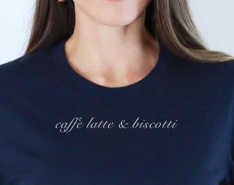May include: Navy blue t-shirt with the text "caffè latte & biscotti" in a delicate, cursive font. The shirt has a classic crew neck and short sleeves. The model is wearing blue jeans and a brown belt.