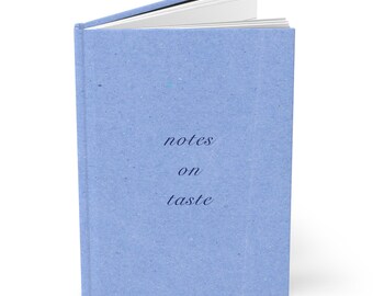 May include: A light blue hardcover notebook with a textured finish. The front cover features the dark blue text "notes on taste" in a serif font. The notebook is slightly open, revealing white pages inside, and stands upright on its spine.