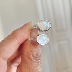 Peut inclure: Une bague en argent avec deux pierres ovales de pierre de lune. Les pierres sont irisées et ont une brillance blanche opalescente.