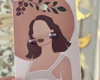 May include: A pair of amethyst stud earrings with silver wire detailing, displayed on a card featuring an illustration of a woman wearing a hat. The earrings are a deep purple color and are round in shape.