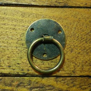 May include: A round, brass-coloured drawer pull with a ring handle. The pull is mounted on a wooden surface.
