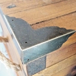 May include: Close-up of a wooden box with a metal corner brace. The brace is a decorative element and has a slightly aged look. The box is made of natural wood with a rustic finish.