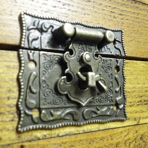 May include: An ornate metal latch with a decorative design, featuring a floral pattern and a small, intricate lock. The latch is made of a dark, aged metal and has a slightly textured surface.