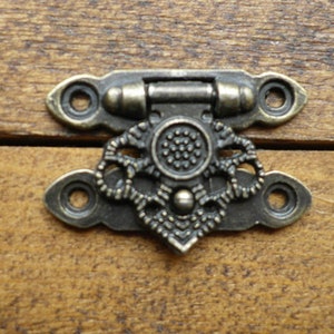 May include: A bronze-coloured metal latch with a decorative design. The latch has two holes on each side and a small knob in the centre.