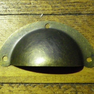 May include: An antique brass drawer pull, shaped like a half-moon, with three mounting holes. The hardware has a curved design and a dark, aged finish. The pull is set against a wooden background.