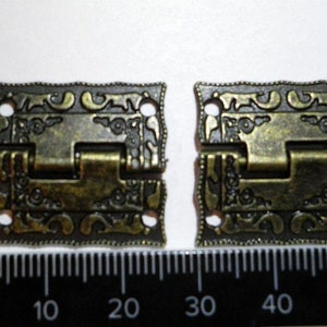May include: Two antique gold-toned metal hinges with ornate designs. The hinges are approximately 5 cm in length and have a decorative floral pattern.
