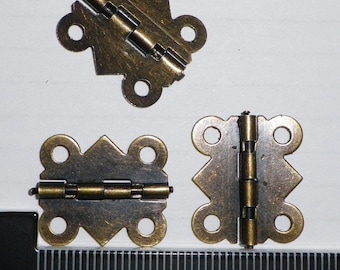 May include: Three antique brass-colored hinges with a decorative diamond and circle design. Each hinge has a central folding mechanism and four circular mounting holes. The hinges are shown against a white background, with a ruler for scale.