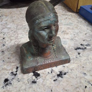 May include: Bronze bust of a Native American woman with a feather in her hair. The bust is mounted on a rectangular base with the name "ANNIE COSTA" inscribed. The sculpture has a weathered, aged appearance.