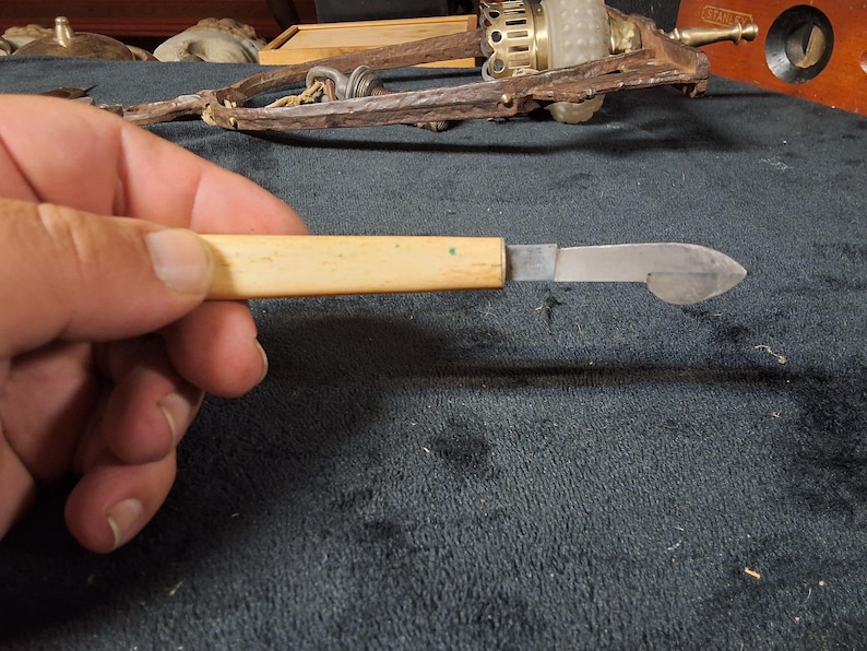 May include: A small, vintage knife with a light-colored handle and a metal blade. The blade has a curved, pointed end. The knife is held in a hand against a dark background. Other antique tools are visible in the background.