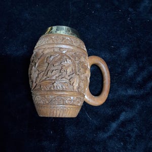 May include: A carved wooden mug with a handle and a brass rim. The mug features detailed leaf carvings around the body. The handle is curved and made of the same wood. The mug is set against a dark background.