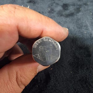 May include: A silver-colored metal object with the words "WEST GERMANY" and a design etched on its surface. The object has a hexagonal shape and appears to be a small, collectible item or component.