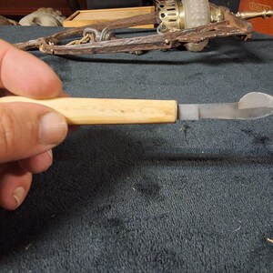 May include: A vintage, small knife with a light-colored wooden handle and a silver-colored blade. The blade has a curved, pointed end. The knife is held in a hand against a dark background.