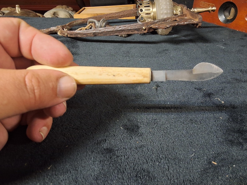 May include: A vintage, small knife with a light-colored wooden handle and a silver-colored blade. The blade has a curved, pointed end. The knife is held in a hand against a dark background.