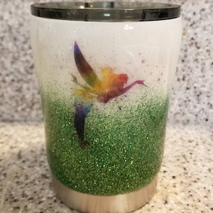 May include: A white tumbler with a green glitter bottom and a rainbow fairy design. The fairy is flying with a wand and stars are scattered around it.