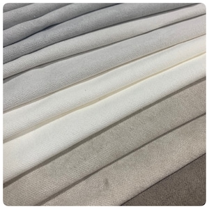 May include: A close-up of a stack of fabric swatches in various shades of white and grey. The fabric has a soft, textured surface.