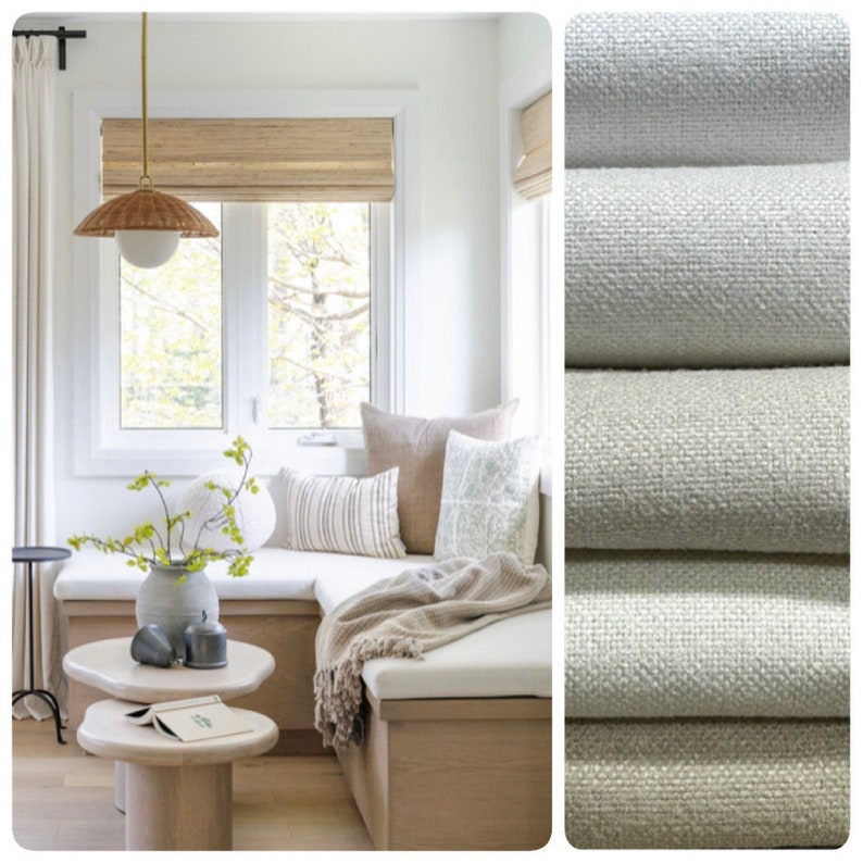 May include: A white window seat with a light brown wooden frame and a white cushion. The seat is decorated with a brown throw blanket and several pillows. The window has a light brown woven shade and a view of trees outside. The seat is in a room with white walls and a light brown wooden floor.