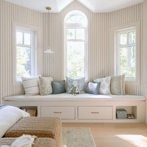 May include: A white window seat with a cushioned bench and storage drawers. The seat is decorated with pillows in various shades of blue and white. The room has white walls with vertical wood paneling and a light wood floor.