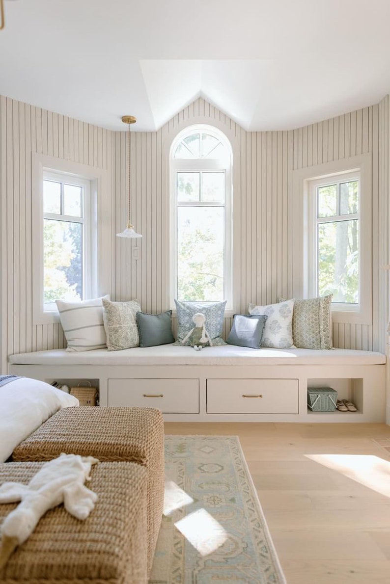 May include: A white window seat with a cushioned bench and storage drawers. The seat is decorated with pillows in various shades of blue and white. The room has white walls with vertical wood paneling and a light wood floor.