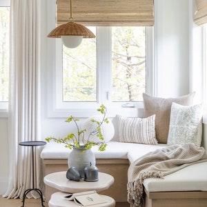 May include: A white window seat with beige cushions and a brown throw blanket. The seat is decorated with throw pillows in various patterns and textures. A small table with a book sits in front of the seat. A vase with a green plant sits on the seat.
