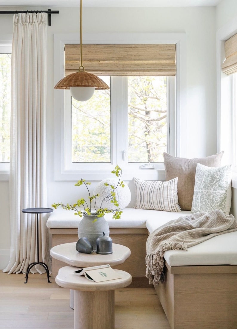 May include: A white window seat with beige cushions and a brown throw blanket. The seat is decorated with throw pillows in various patterns and textures. A small table with a book sits in front of the seat. A vase with a green plant sits on the seat.