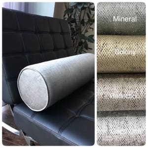 May include: A grey cylindrical pillow on a black leather chair. The pillow is made of a soft, textured fabric. The image also shows four fabric swatches with the names "Mineral", "Colony", "Reflection", and "Graphite".