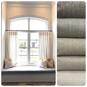 May include: A window seat with a white cushion and two pillows. The window has a curved top and is covered with white curtains with a pattern of small circles. The curtains are hung on a silver rod. The window seat is in front of a large window with a view of a house and trees. The image also shows a close-up of four different fabric swatches in various shades of gray.