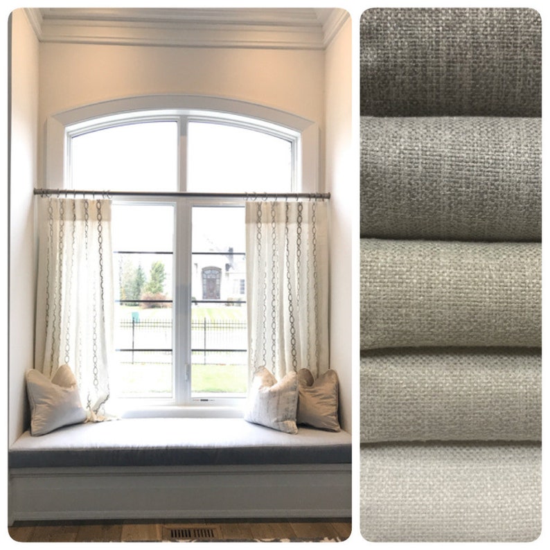May include: A window seat with a white cushion and two pillows. The window has a curved top and is covered with white curtains with a pattern of small circles. The curtains are hung on a silver rod. The window seat is in front of a large window with a view of a house and trees. The image also shows a close-up of four different fabric swatches in various shades of gray.