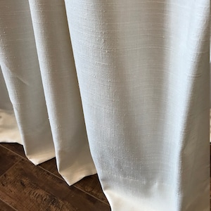 May include: Close-up of white linen curtains with a subtle texture. The curtains are gathered at the bottom and hang against a wood floor.