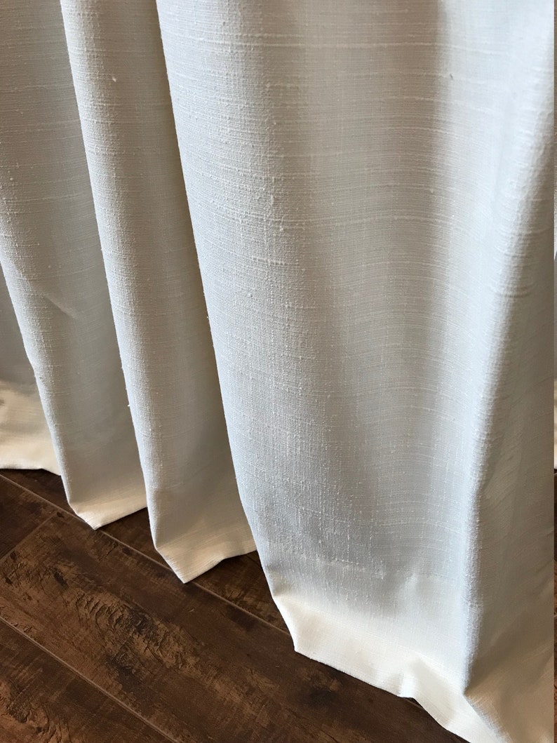 May include: Close-up of white linen curtains with a subtle texture. The curtains are gathered at the bottom and hang against a wood floor.