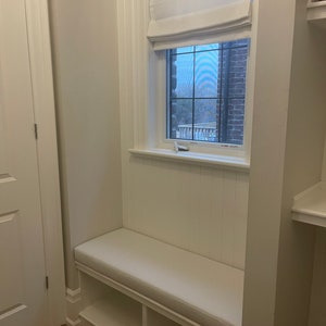 Puede incluir: Asiento de ventana blanco con un coj&iacute;n y dos estantes debajo. La ventana tiene una cortina romana blanca.