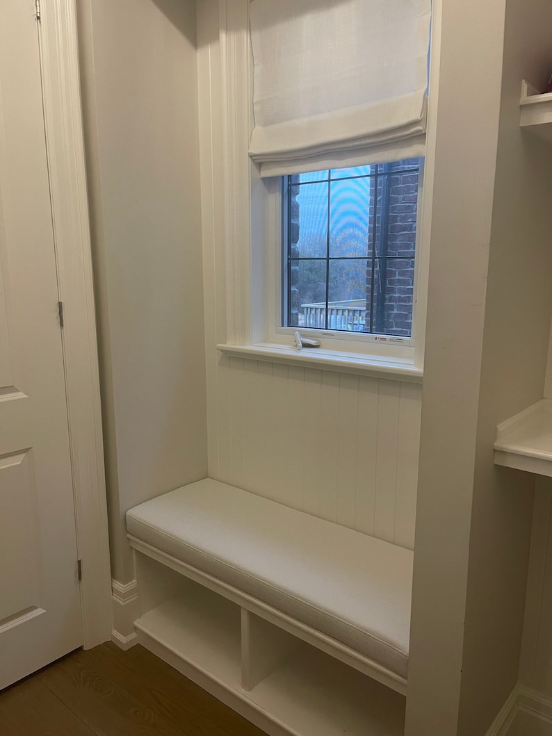 Puede incluir: Asiento de ventana blanco con un coj&iacute;n y dos estantes debajo. La ventana tiene una cortina romana blanca.