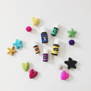 May include: A collection of essential oil bottles in various colors, including blue, green, and yellow, arranged on a white surface with felt hearts, stars, and circles in different colors.
