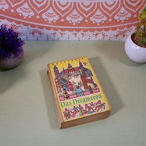 May include: A vintage copy of "Das Decameron" by Boccaccio. The book has a yellow cover with a colourful illustration of a city scene. The title is in a teal font. The book is resting on a light green surface.