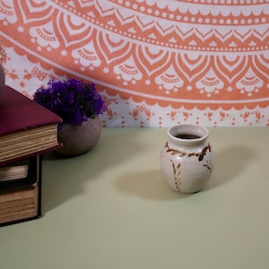 May include: A small, white ceramic vase with brown painted designs. The vase is sitting on a light green surface.